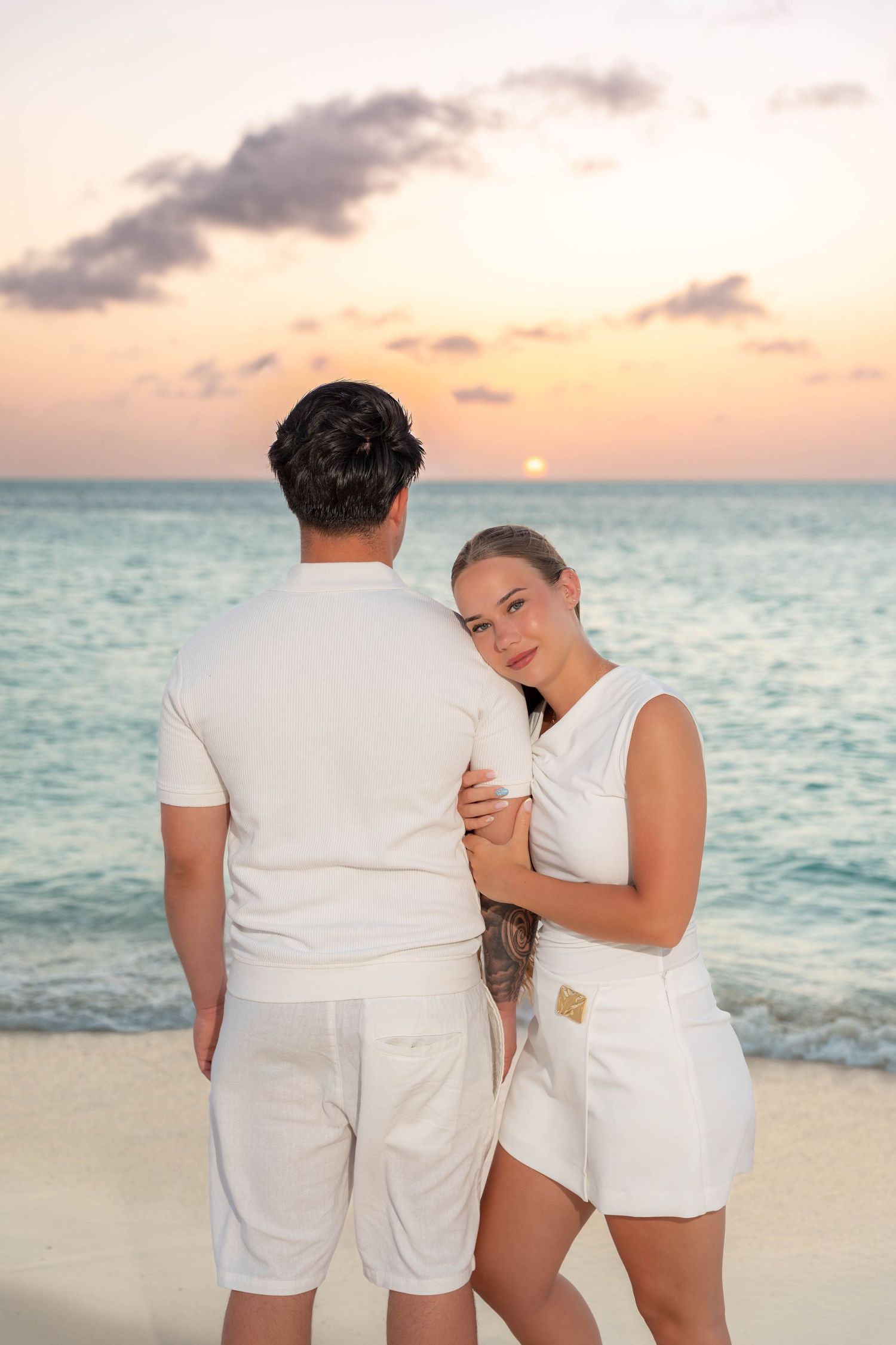 Couple in white outfits embracing on a sandy Aruba beach at sunset with turquoise ocean waves in the background – Jonathan Geerman Studio