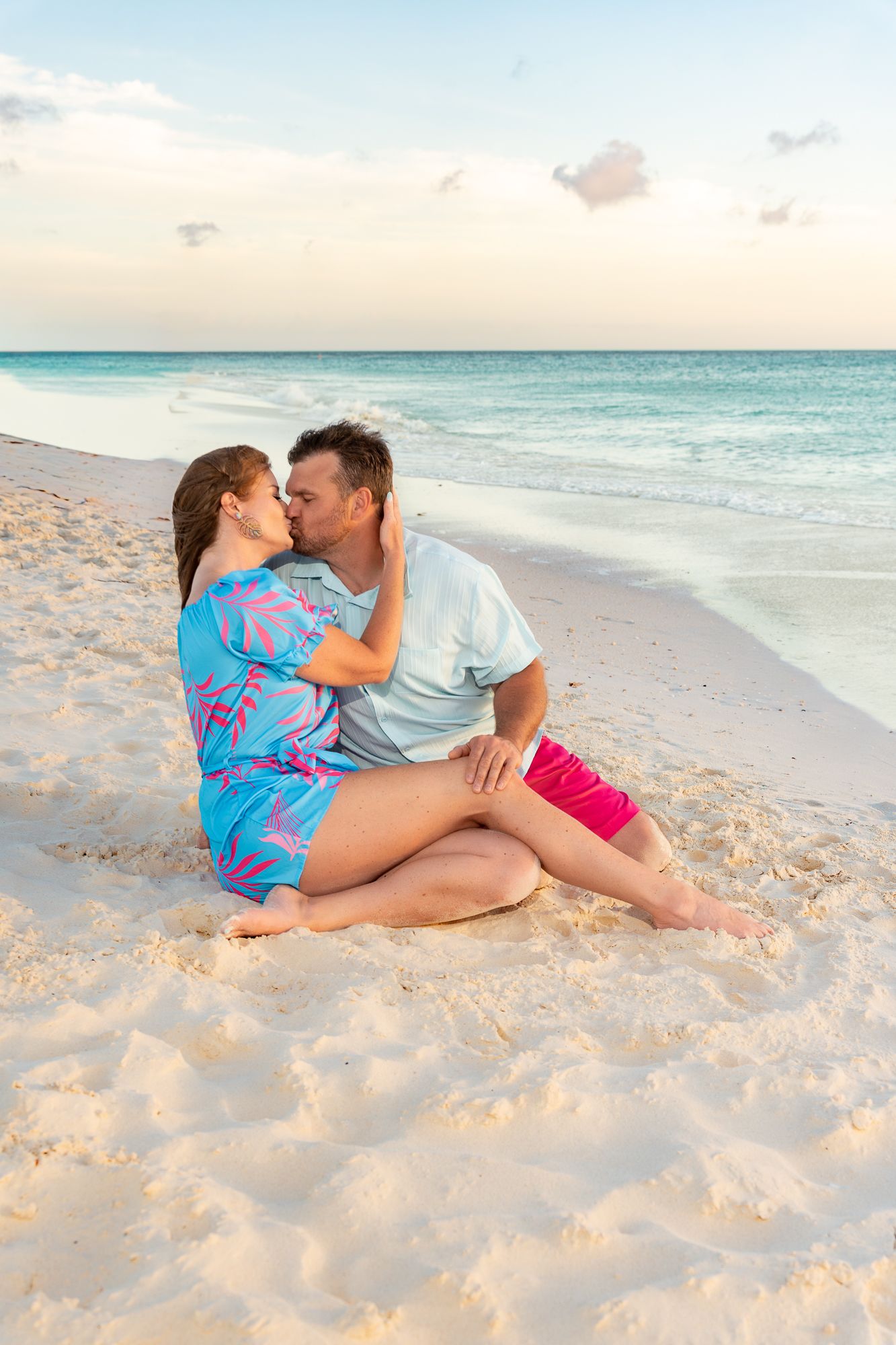 Romantic couple kissing while sitting on white sand beach with turquoise ocean waves at sunset.