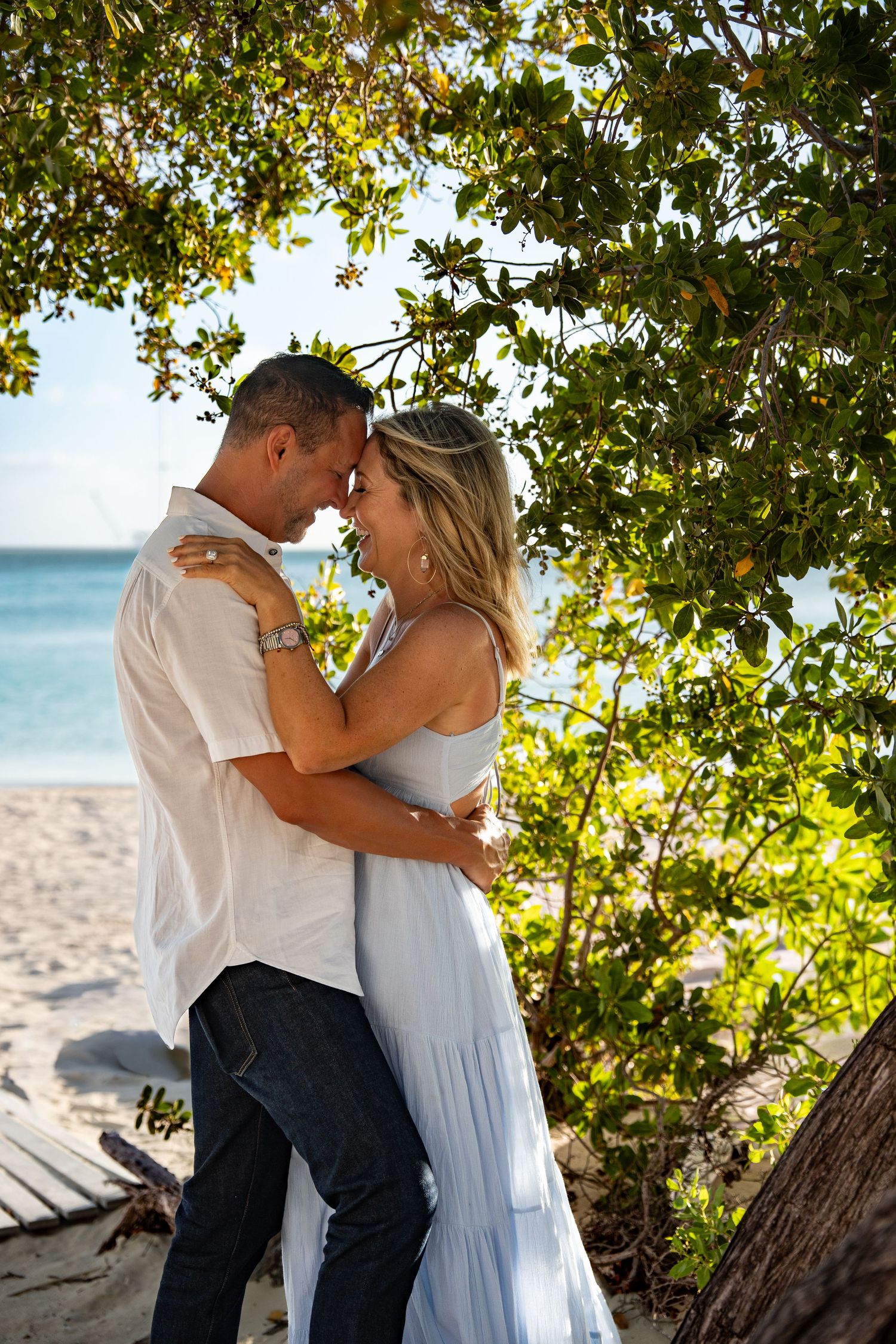 Romantic couple embracing under a lush green tree on a tropical white sand beach.