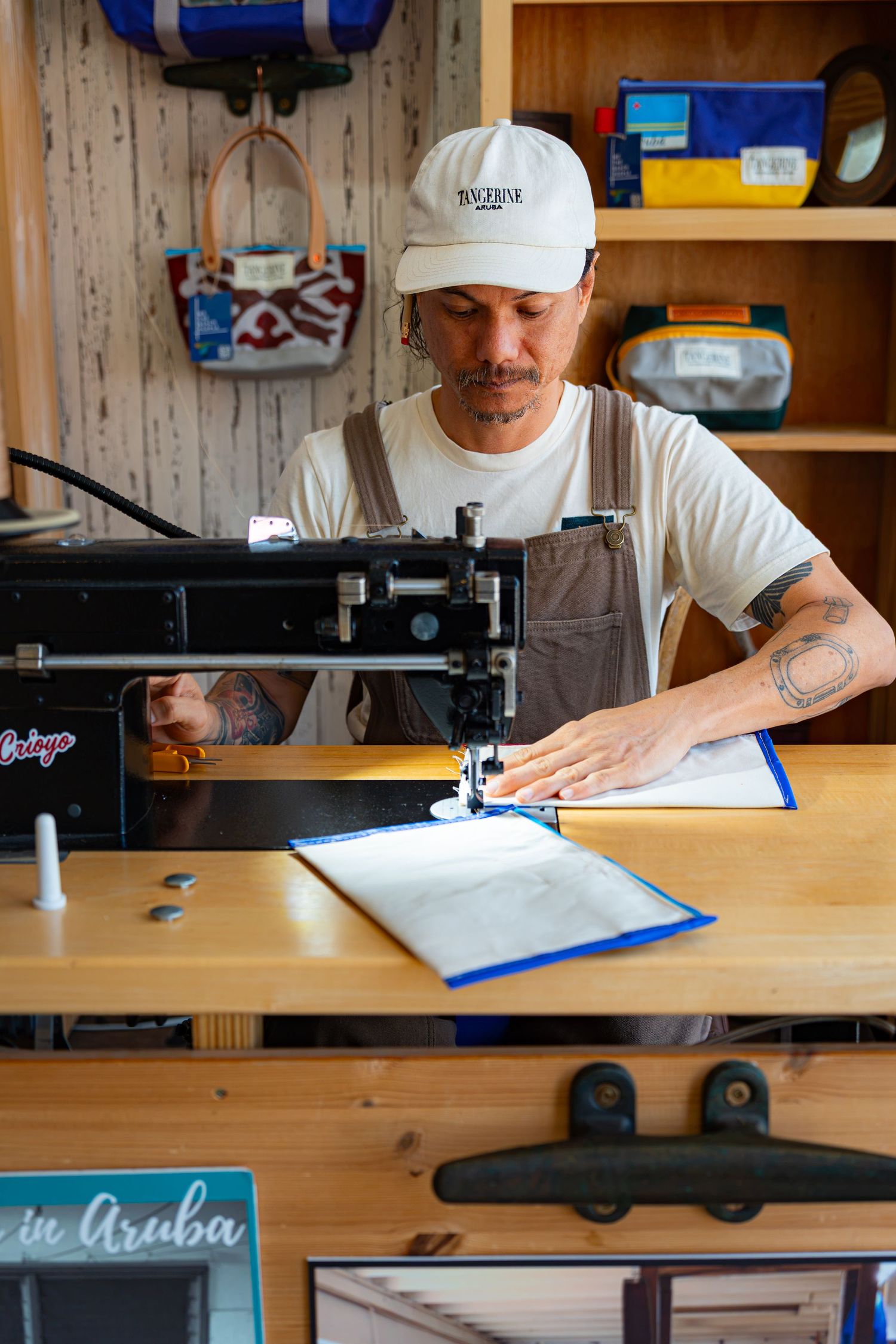 Tattooed craftsman in white cap using sewing machine in a cozy wooden workshop.