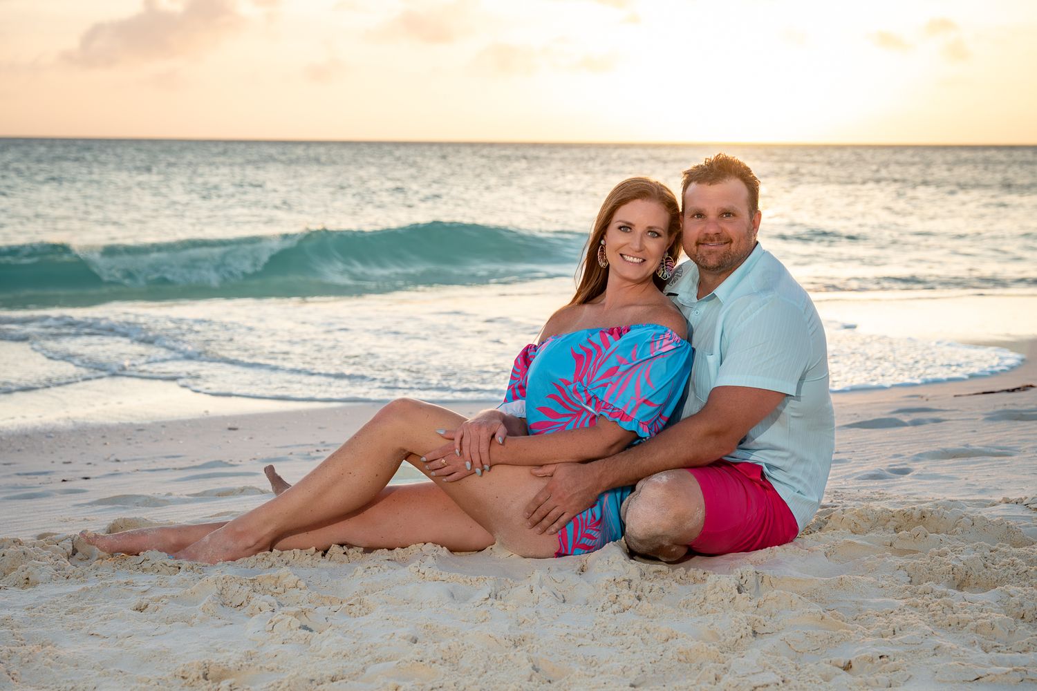 Couple sitting together on a sandy beach at sunset, smiling warmly at the camera.