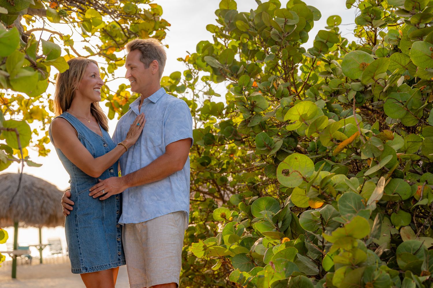 Smiling couple embracing romantically among lush green tropical plants on a sunny beach.