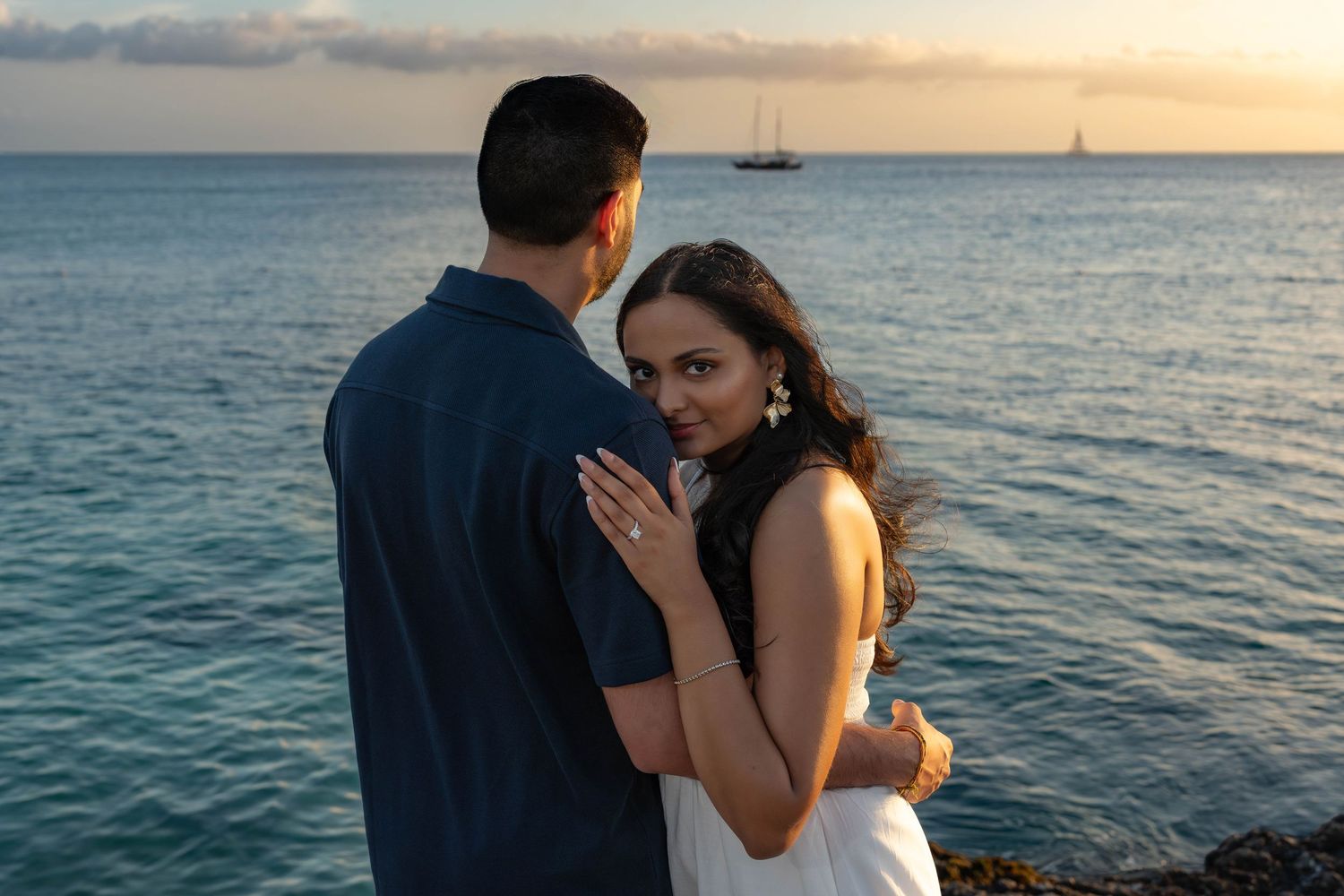 Woman in white dress embracing man near a rocky ocean shore at golden sunset in Aruba – Jonathan Geerman Studio