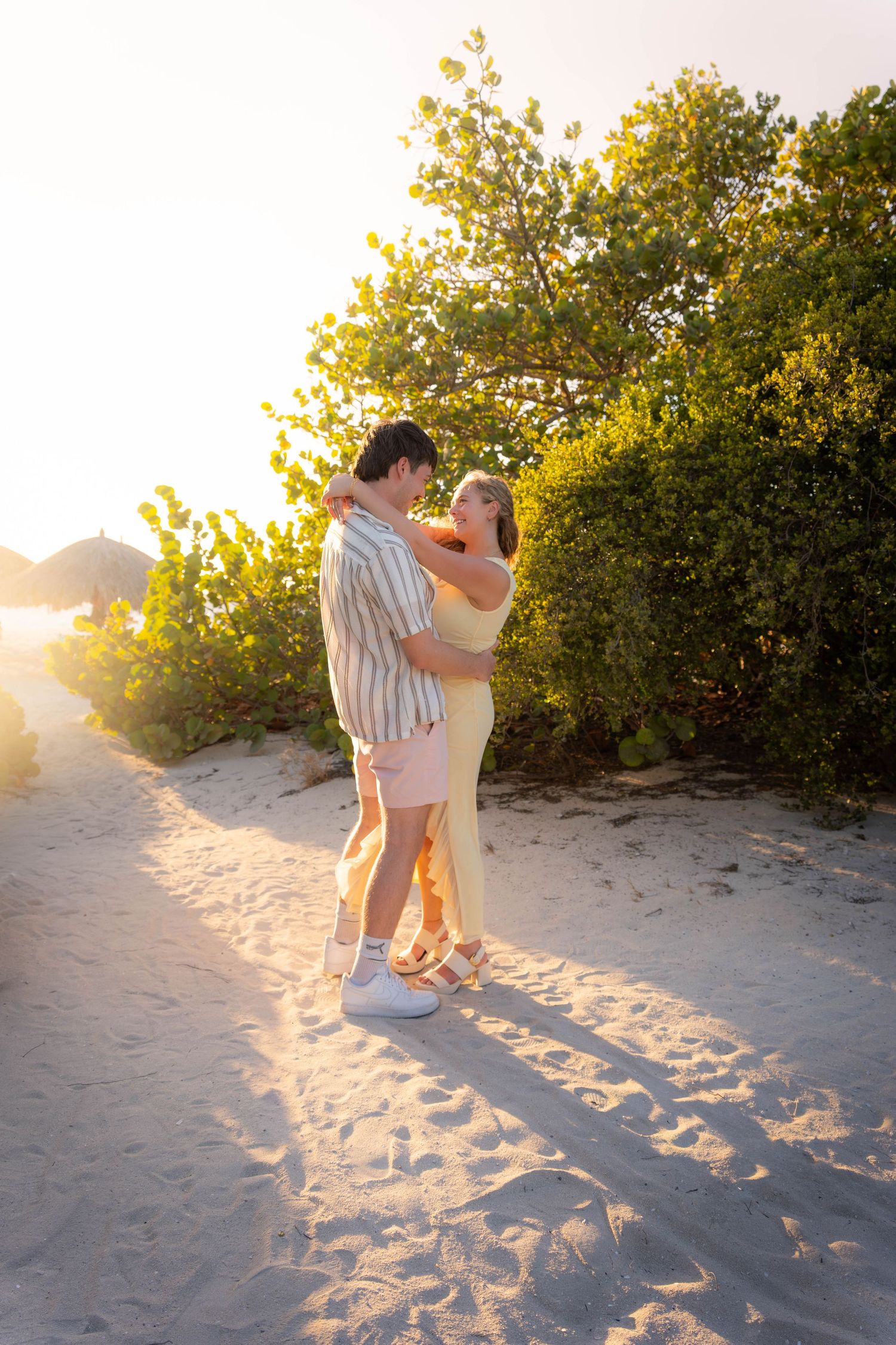 Romantic couple embracing on a sandy beach path at golden sunset, surrounded by lush green tropical foliage.