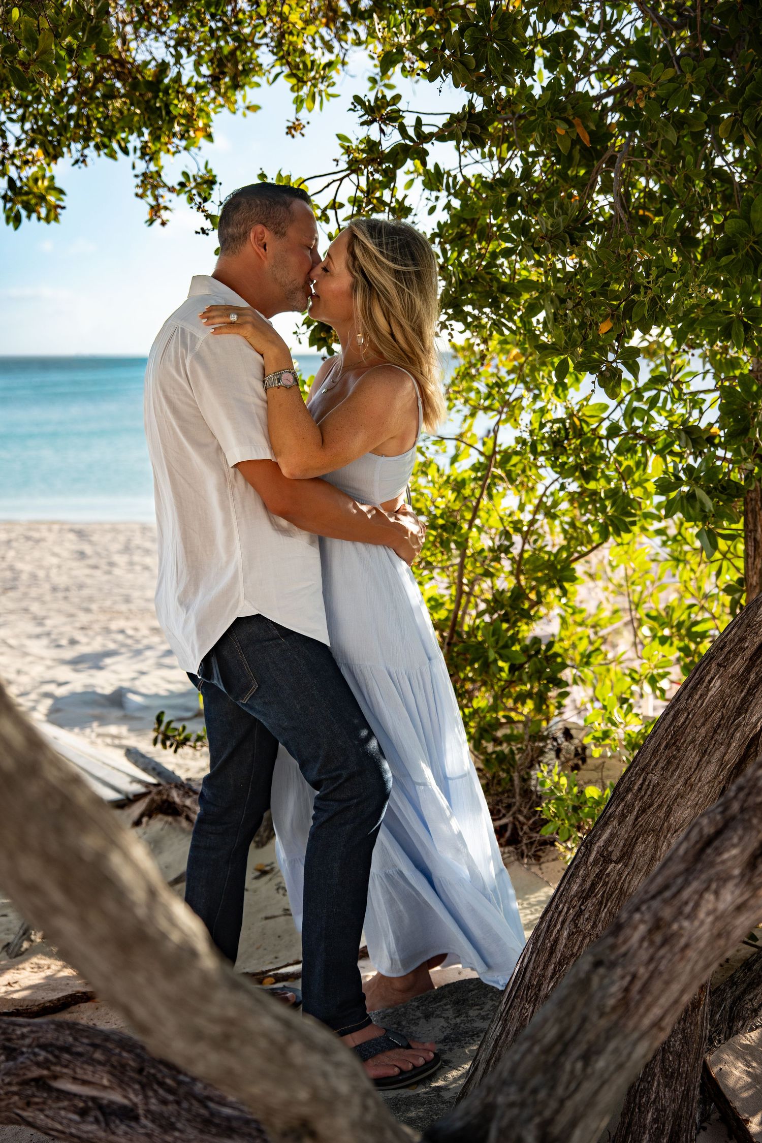 Couple kissing and embracing under lush green trees on a sandy tropical beach.