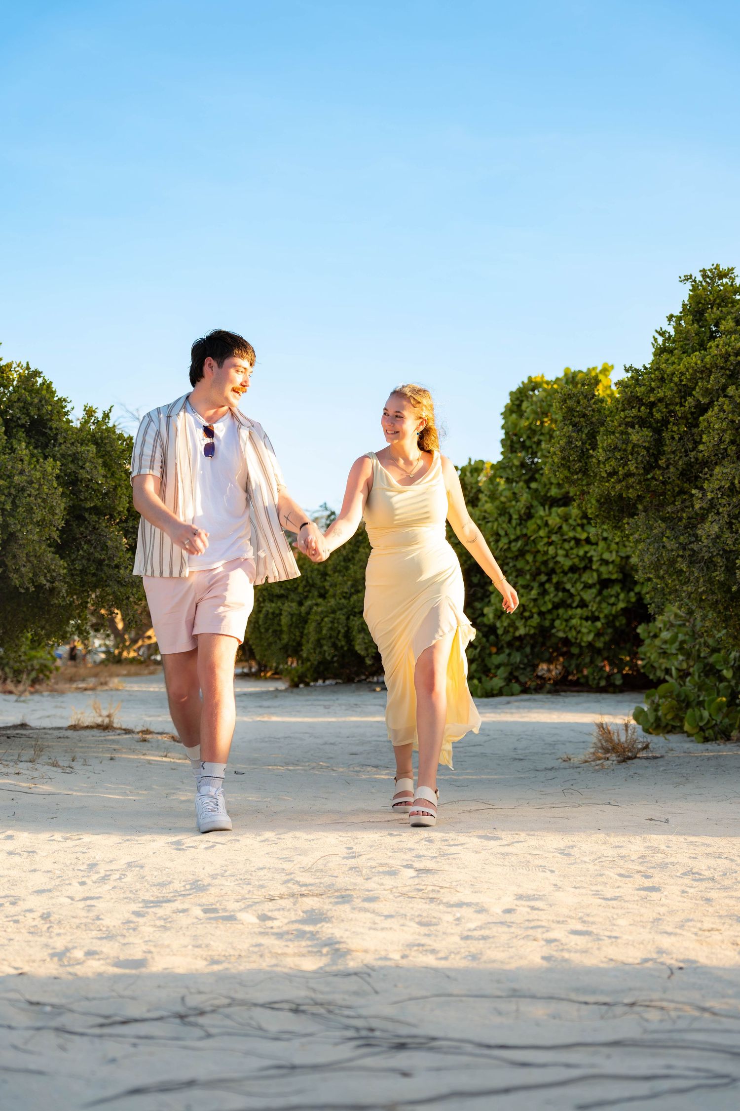 Young couple holding hands walking through a sandy beach path in Aruba surrounded by lush tropical greenery – Jonathan Geerman Studio