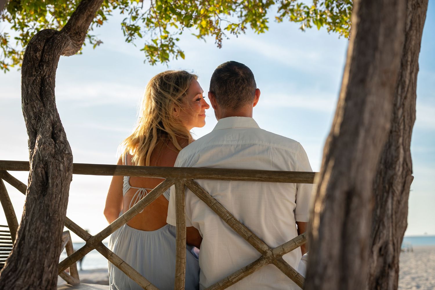 Romantic couple sitting on wooden bench facing each other at tropical beach sunset.