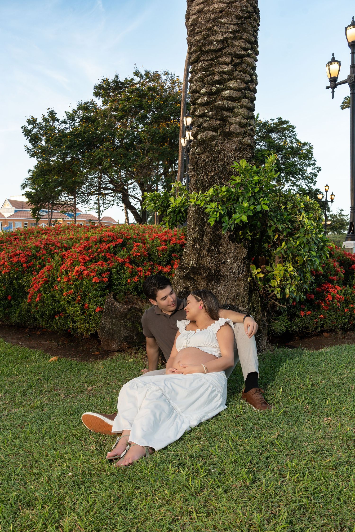 Pregnant couple sitting together against palm tree surrounded by red flowers at dusk.