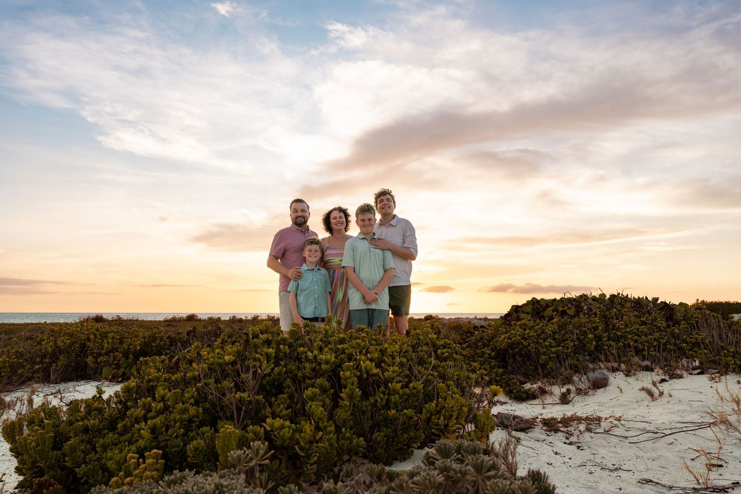 Happy family of five standing together in coastal dunes at sunset in Aruba with a colorful sky – Jonathan Geerman Studio