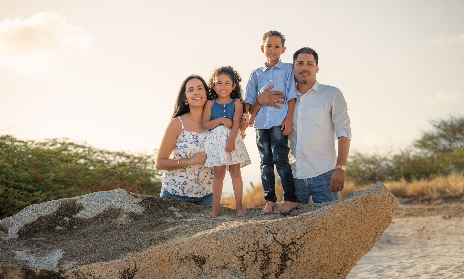 Family of four posing on a large rock at golden hour in Aruba with warm sky and lush greenery behind them – Jonathan Geerman Studio