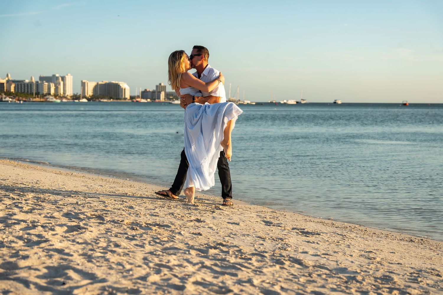 Couple kissing as man lifts woman in white dress on sunny tropical beach.
