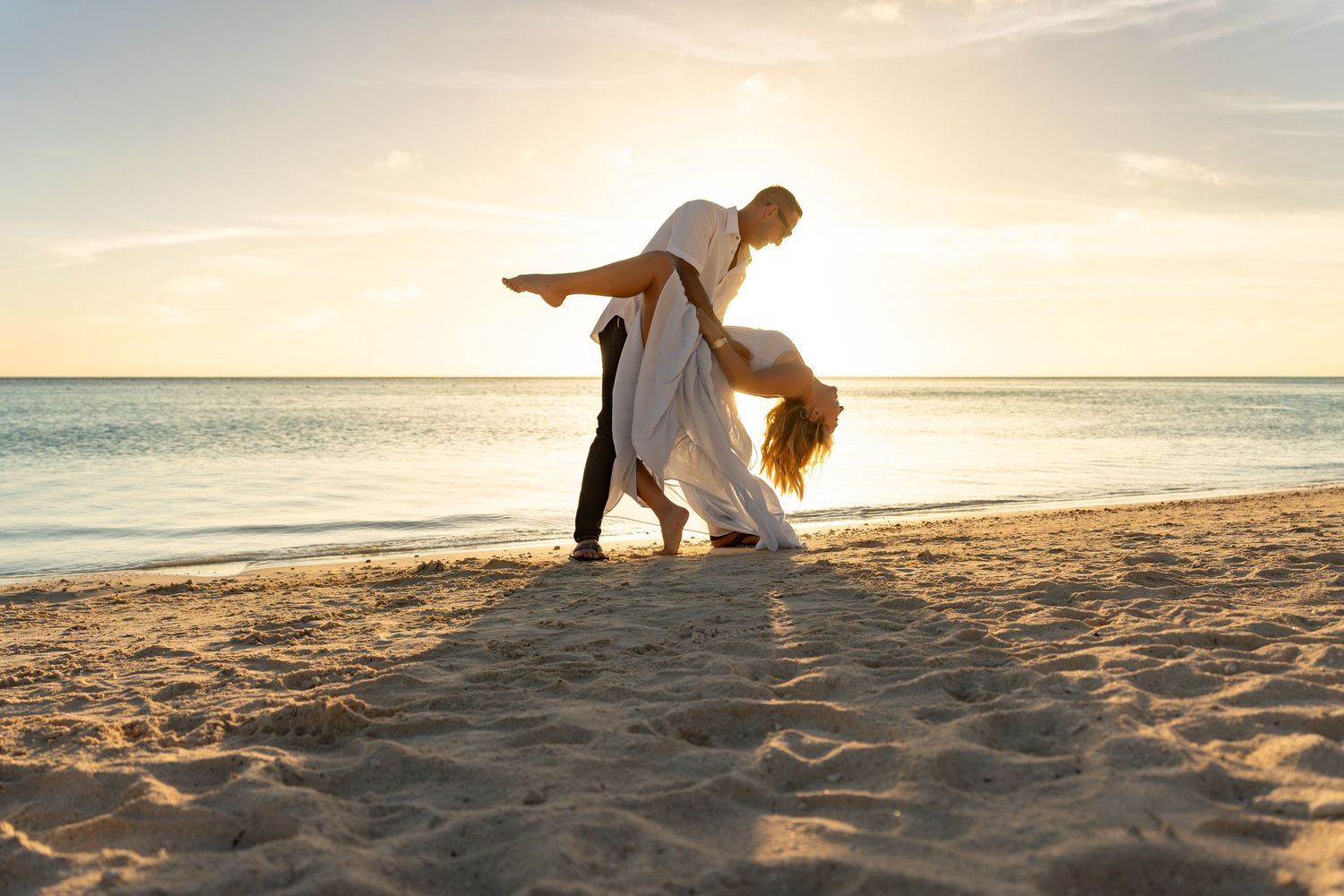 Couple dancing romantically on a sandy beach at sunset, man dipping woman in white dress by the ocean.