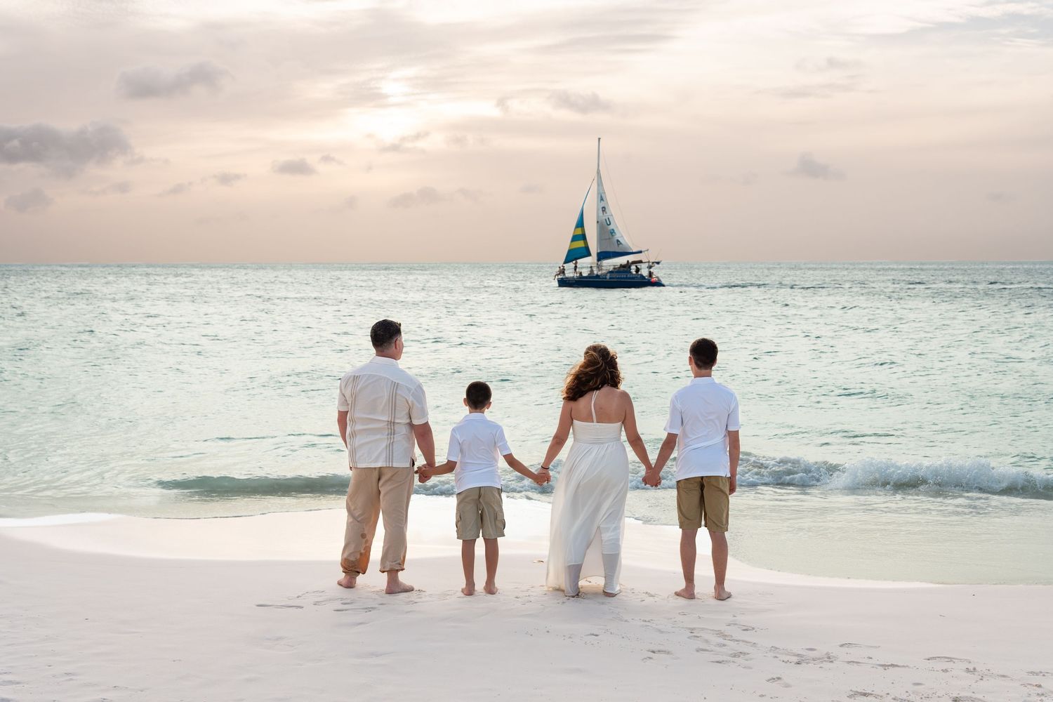 Family of five holding hands on an Aruba beach at sunset, watching a sailboat, backs to the camera.