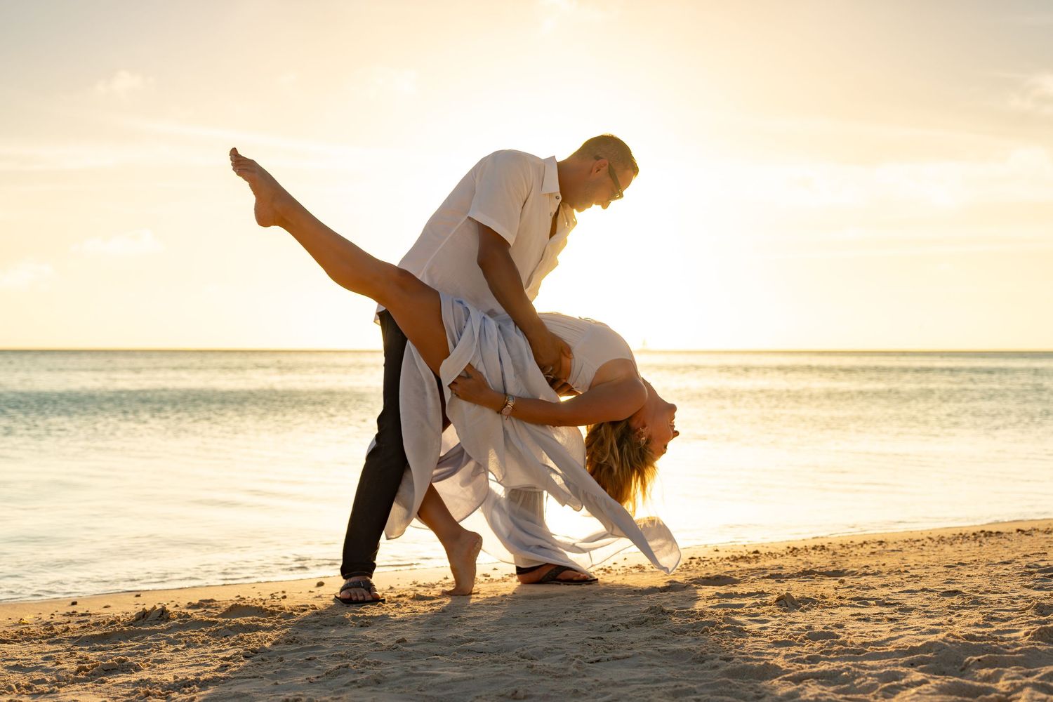 Man dipping woman dramatically on sandy beach during golden sunset by the ocean.