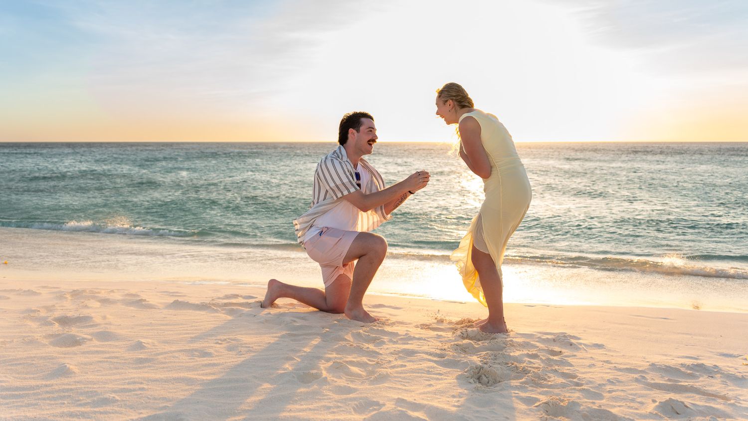 Man kneeling to propose to a surprised woman on an Aruba beach at golden hour, surprise proposal photoshoot