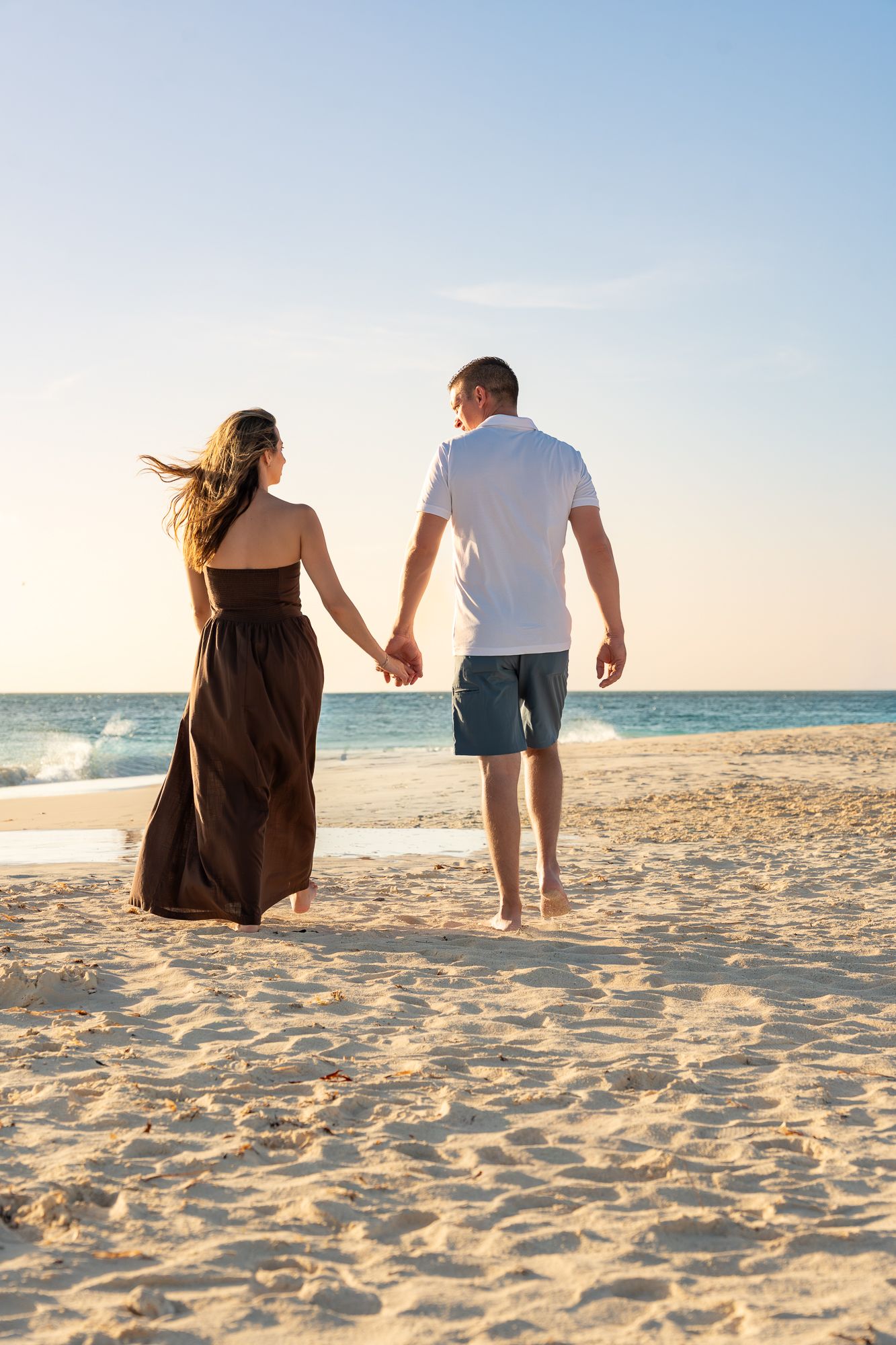 Couple holding hands walking toward the ocean on a sandy beach at golden sunset.