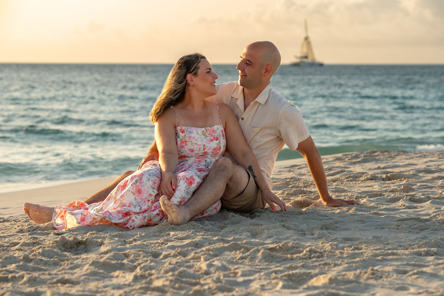 A couple sits together on a sandy beach at sunset, smiling at each other with a sailboat visible on the ocean.