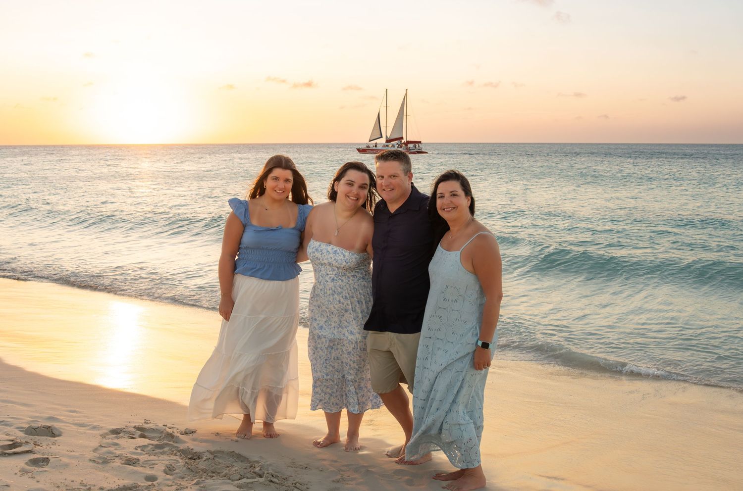 Family of four smiling together on an Aruba beach at sunset with a sailboat visible on the horizon – Jonathan Geerman Studio