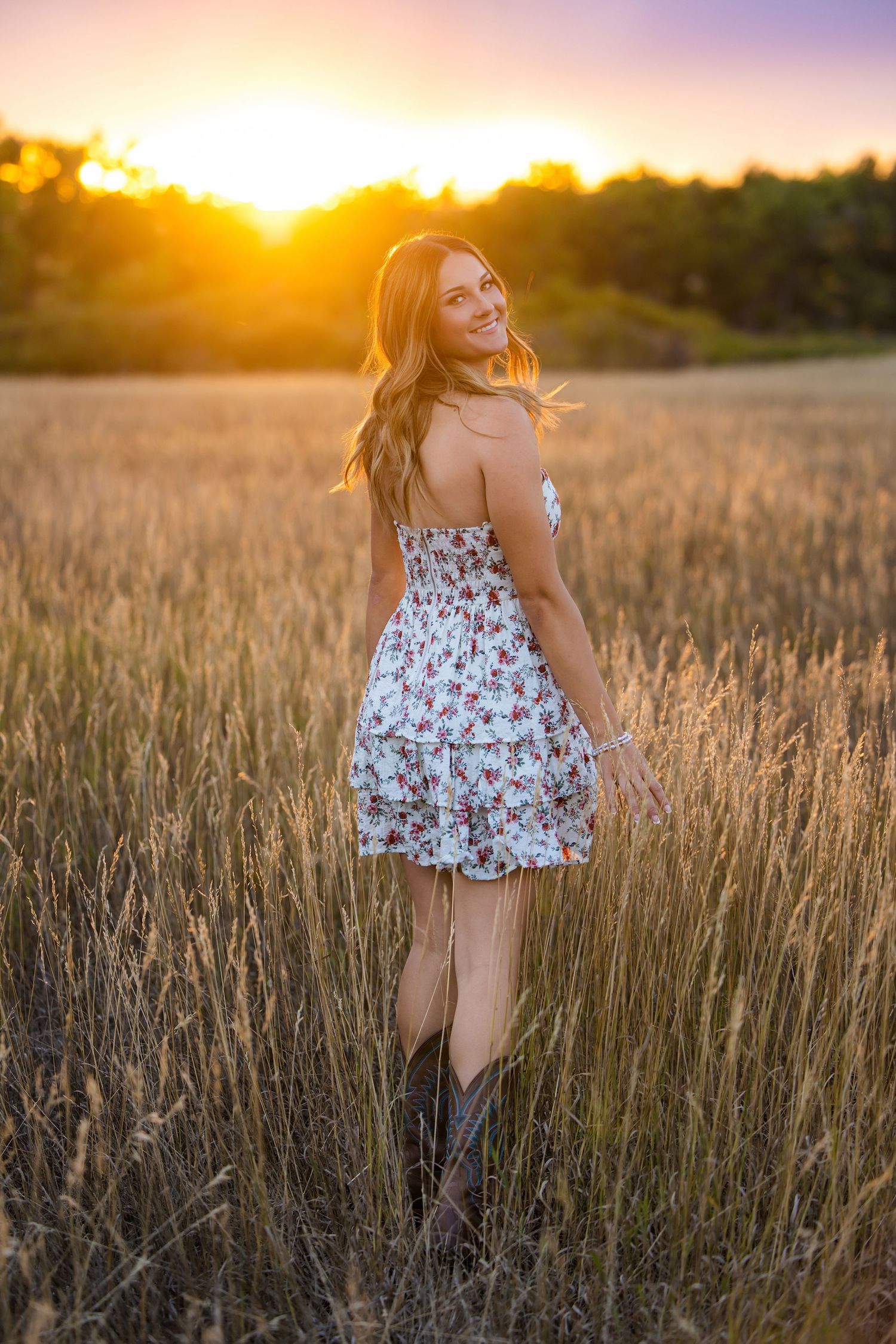 Young woman in floral dress and cowboy boots standing in wheat field during stunning golden sunset.  Parker Colorado Senior Portrait