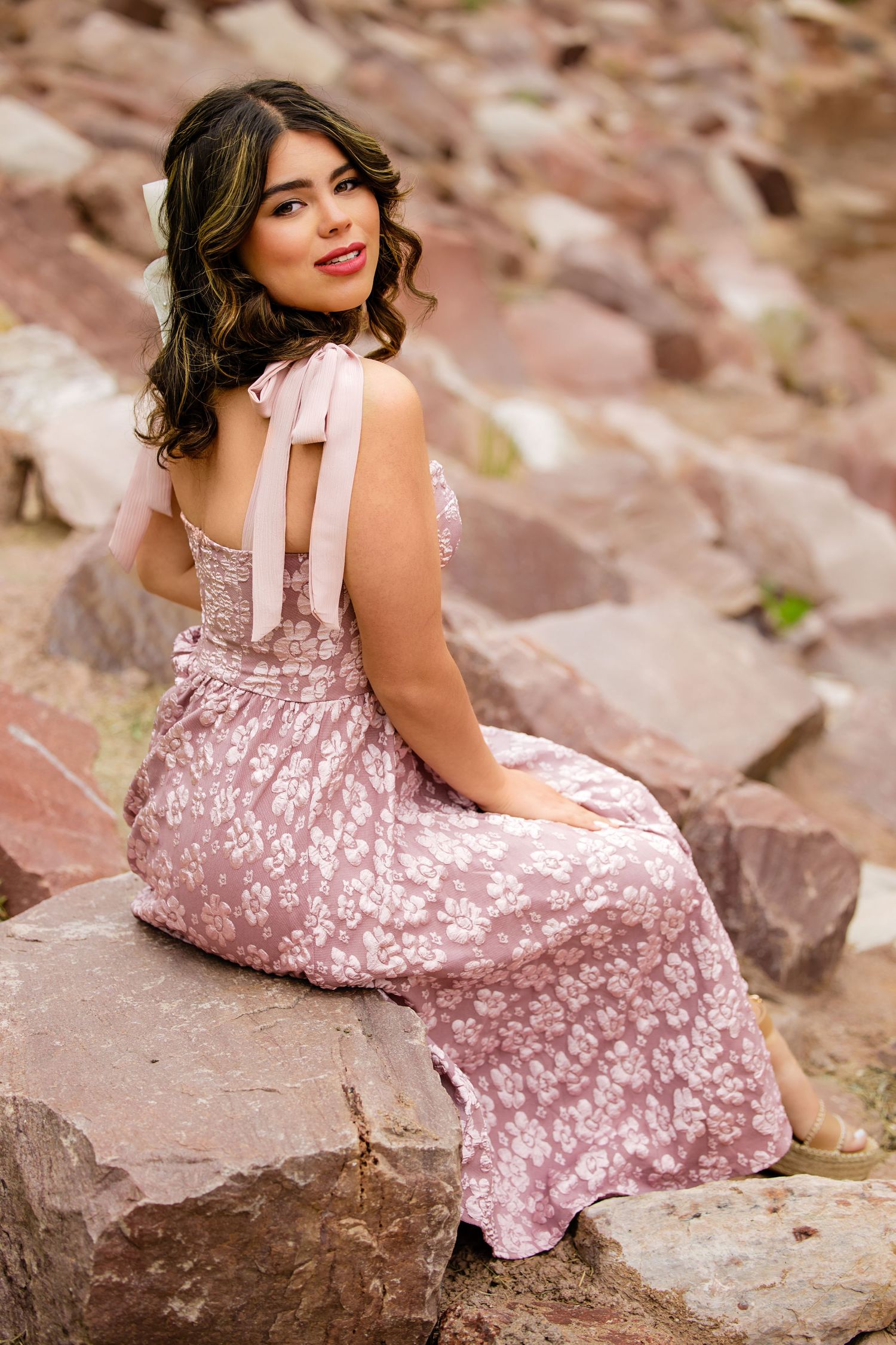 Young woman in pink floral dress with bow back sits on red rocks smiling over her shoulder for Parker Colorado Senior pictures