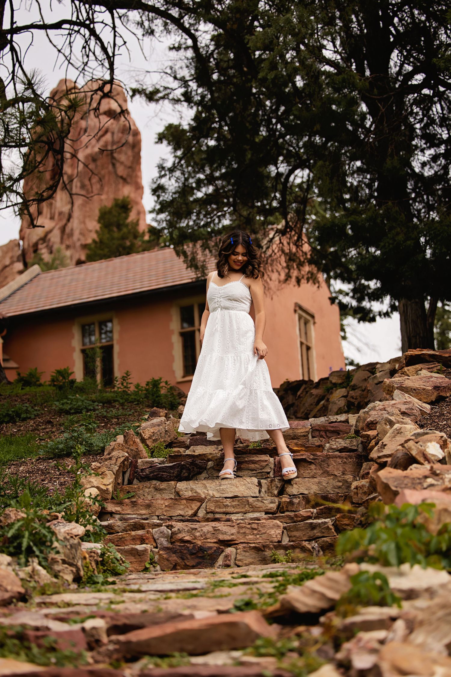 Young woman in white midi dress walking down stone steps near rustic building for Franktown Colorado Senior pictures