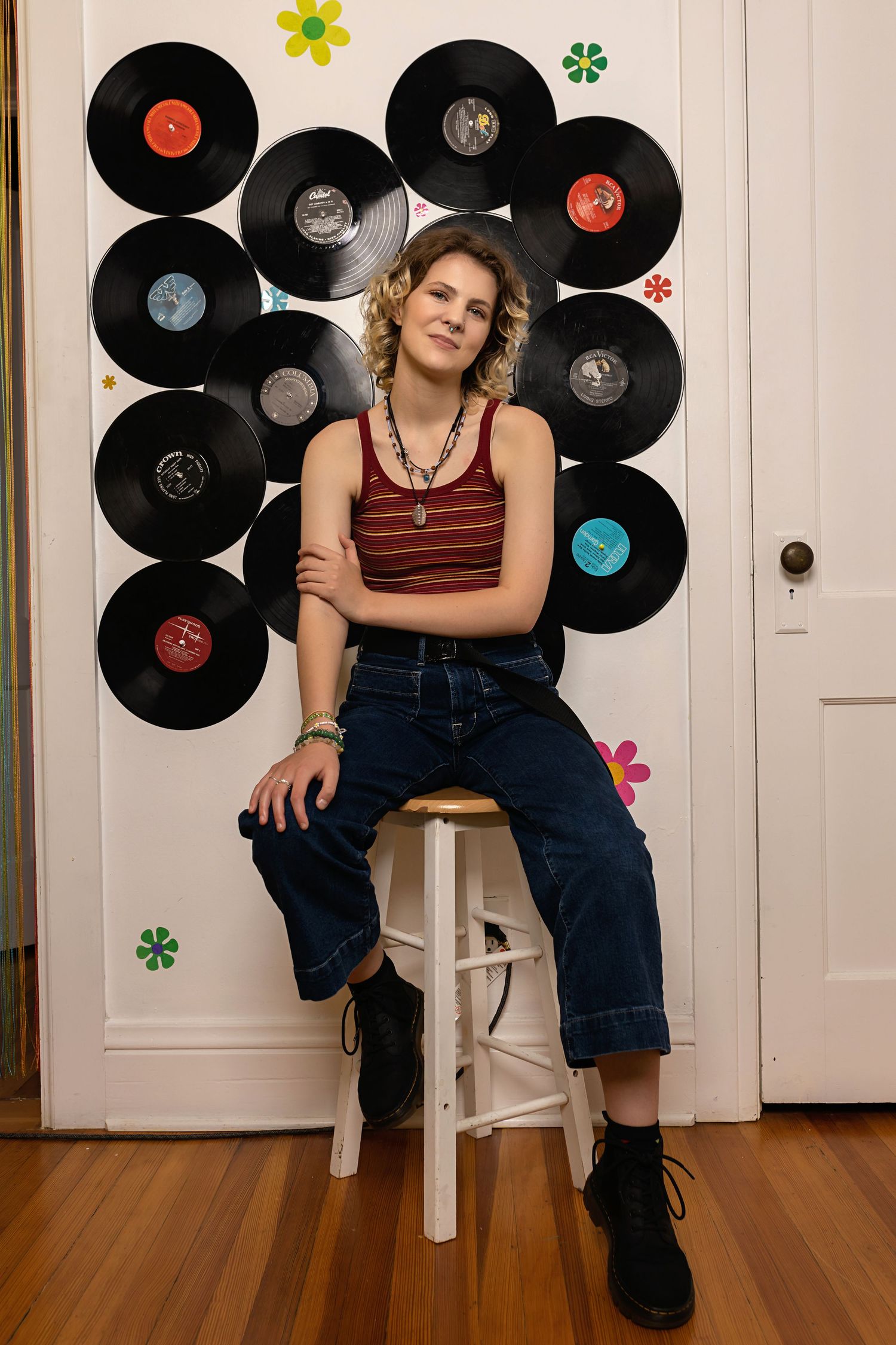 Curly-haired person in striped tank and wide-leg jeans poses before vinyl records on white wall posing for senior photos Parker Colorado