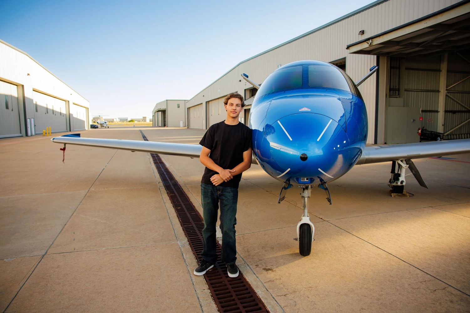 Teenage boy standing next to a blue airplane outside an airport hangar at dusk.