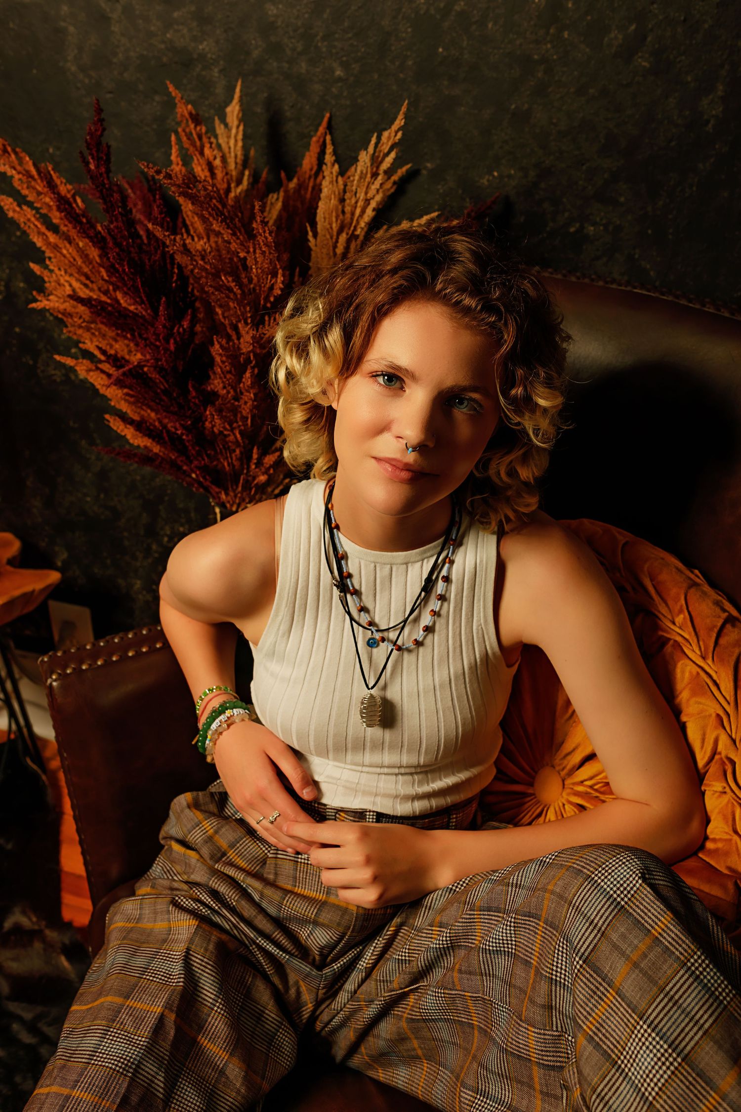 Curly blonde woman in white ribbed tank and plaid pants seated in leather chair with dried botanicals.