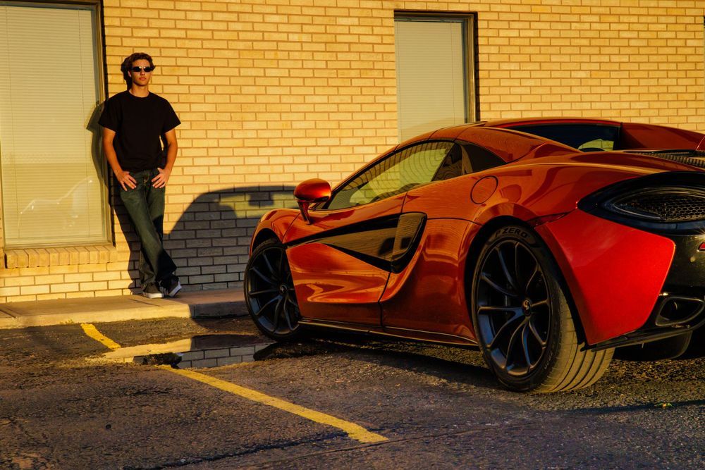 Man in black outfit leaning against brick wall beside a red McLaren sports car in golden hour light.  Colorado Senior Portrait