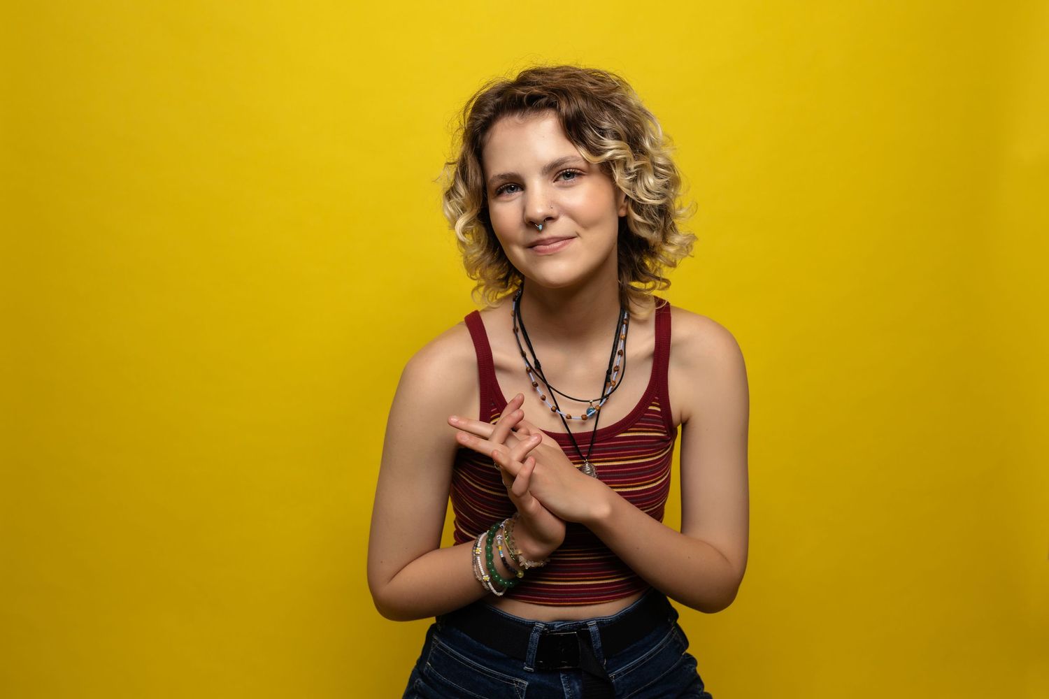 Young person in striped crop top and jeans smiling with hands clasped against bright yellow background. Aurora Colorado Senior Portrait