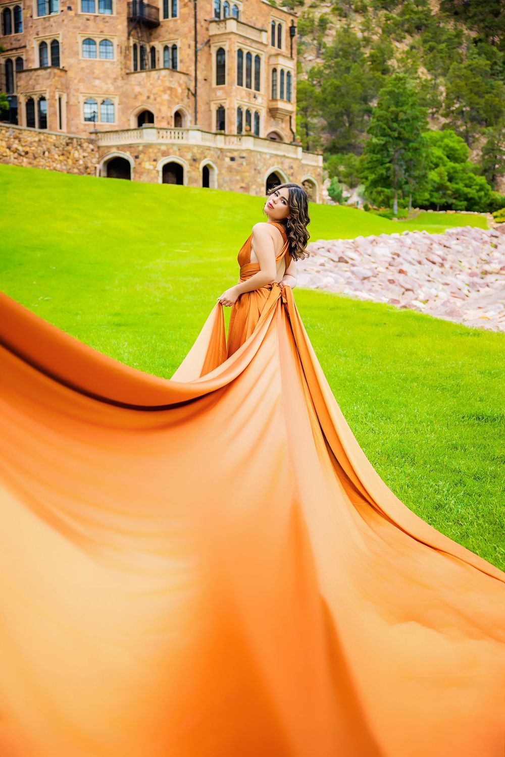 Woman in flowing orange gown with dramatic train billowing in wind before castle on green lawn for senior photos in Colorado