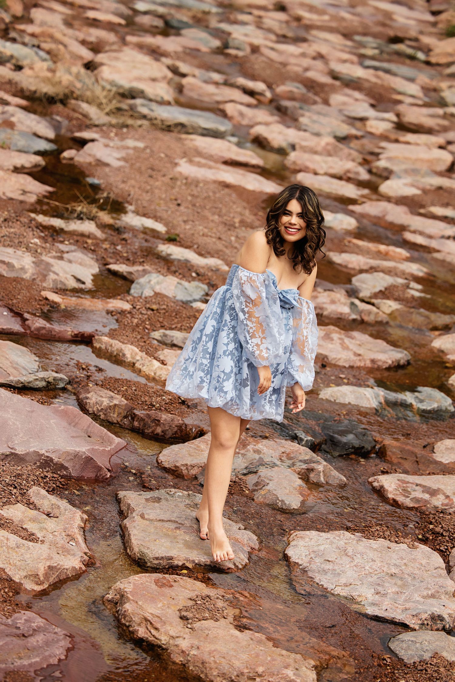 Smiling barefoot woman in blue floral lace dress walking on red rocky terrain outdoors Lonetree CO High School Senior pictures