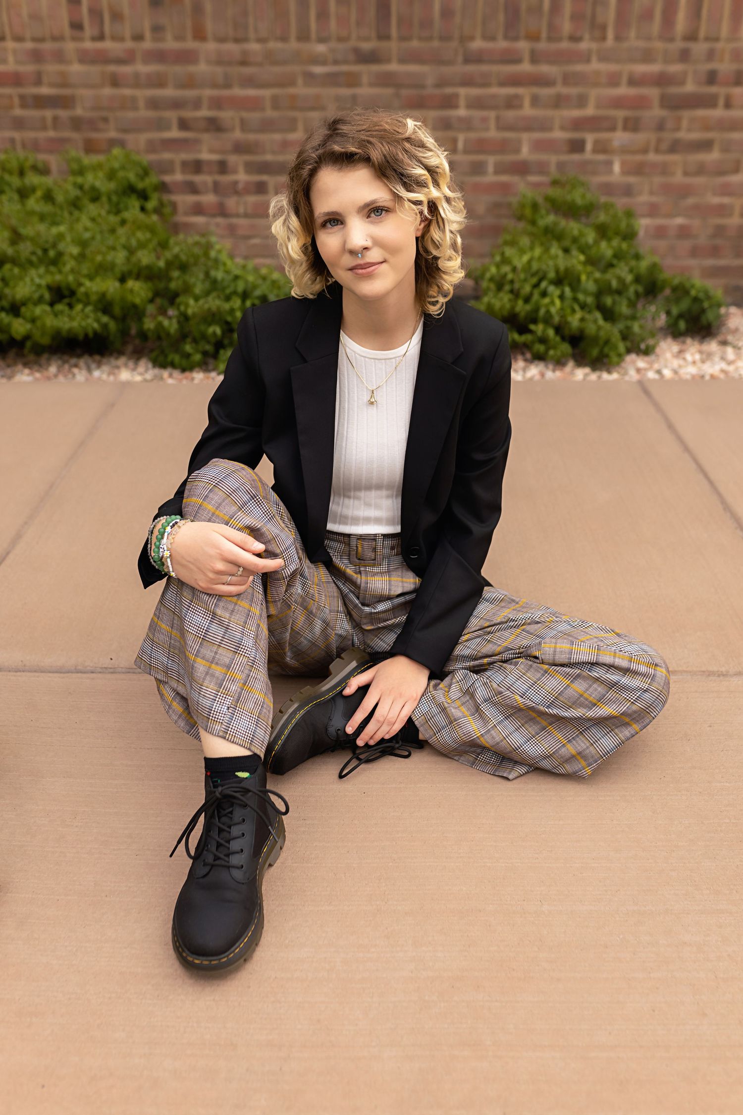 Person in black blazer and plaid pants sits casually on pavement near brick wall. Downtown Denver Senior Photos.