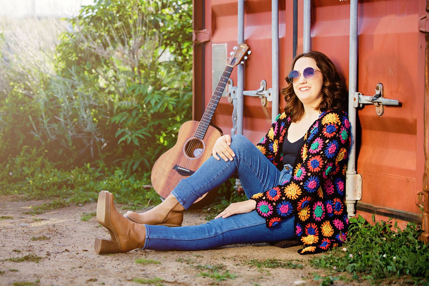 Colorado High School  woman in colorful crochet cardigan and sunglasses sitting outdoors with acoustic guitar leaning against red wall.