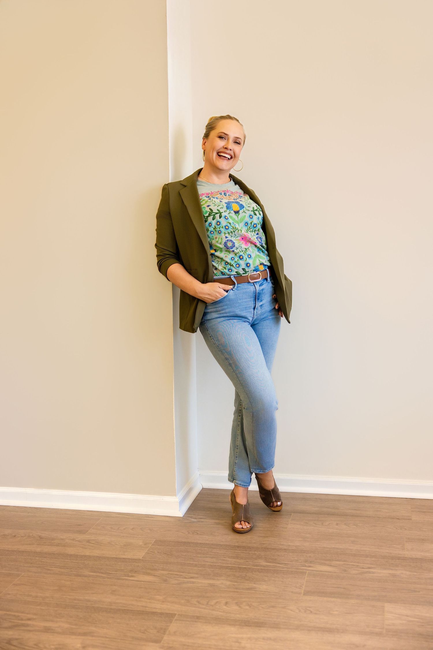 Melissa Nansen: wearing green blazer, floral top, and blue jeans leaning against a wall with wood floors in her Parker Colorado studio