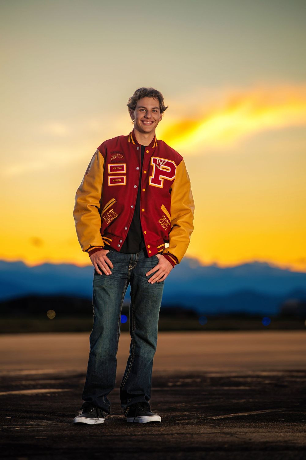 Smiling teen in red gold letterman jacket posing on tarmac with colorful sunset and mountains behind. Classic Colorado Senior Portrait