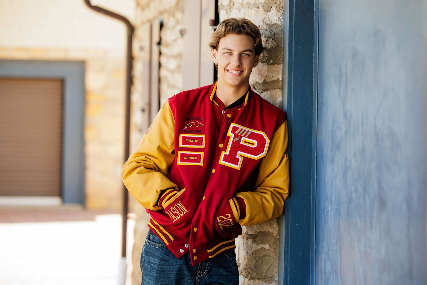 Smiling teen boy in red and gold varsity jacket leaning against a stone wall outdoors. Denver Colorado Senior Portrait