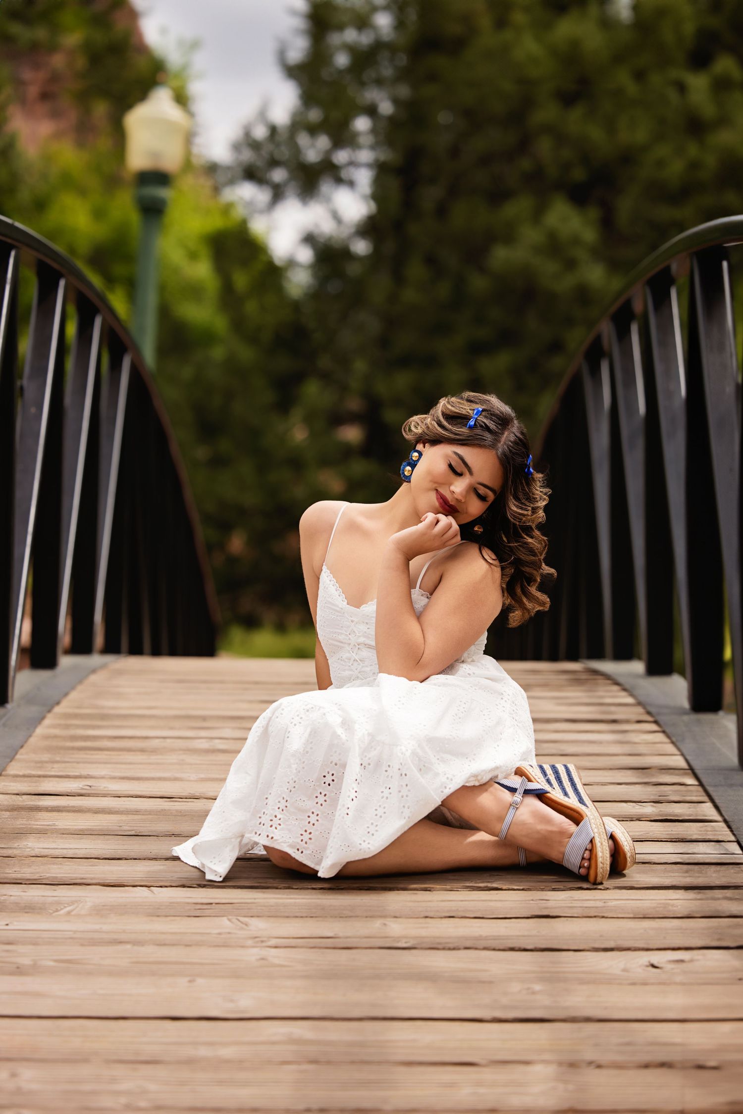 Young woman in white sundress sitting on wooden bridge deck with blue earrings for Parker Colorado Senior pictures