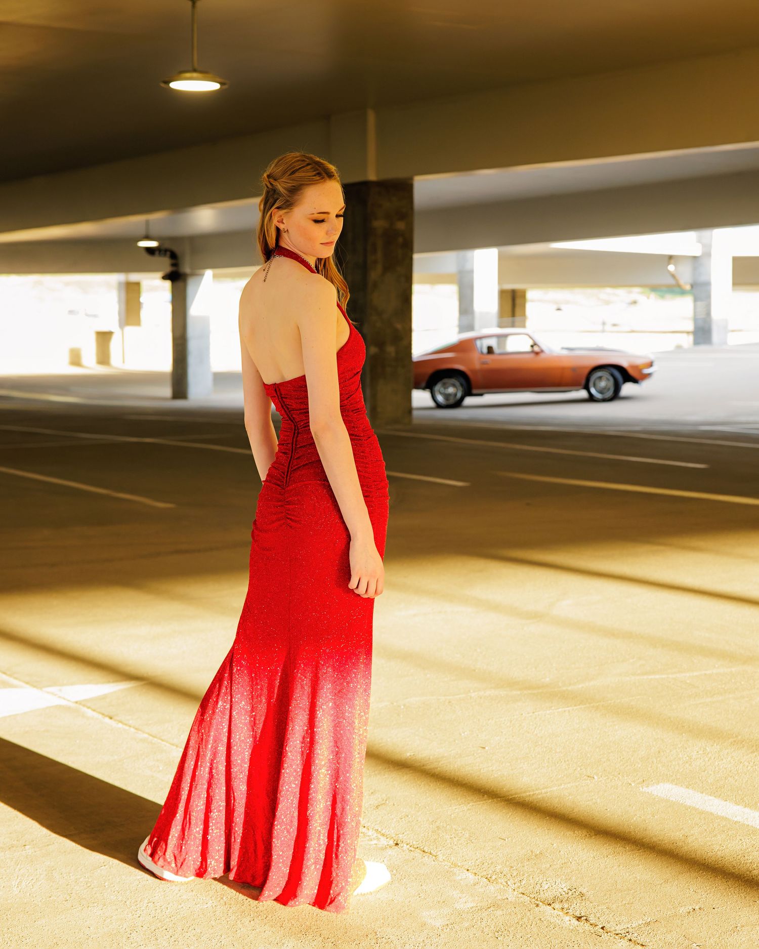 Woman in red halter gown poses in sunlit parking garage with vintage brown car in background in Metro Devnver