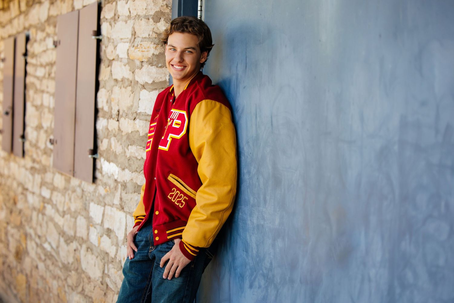 Smiling teen boy in red and gold varsity jacket leaning against blue wall for senior photos. Aurora Colorado Senior Portrait