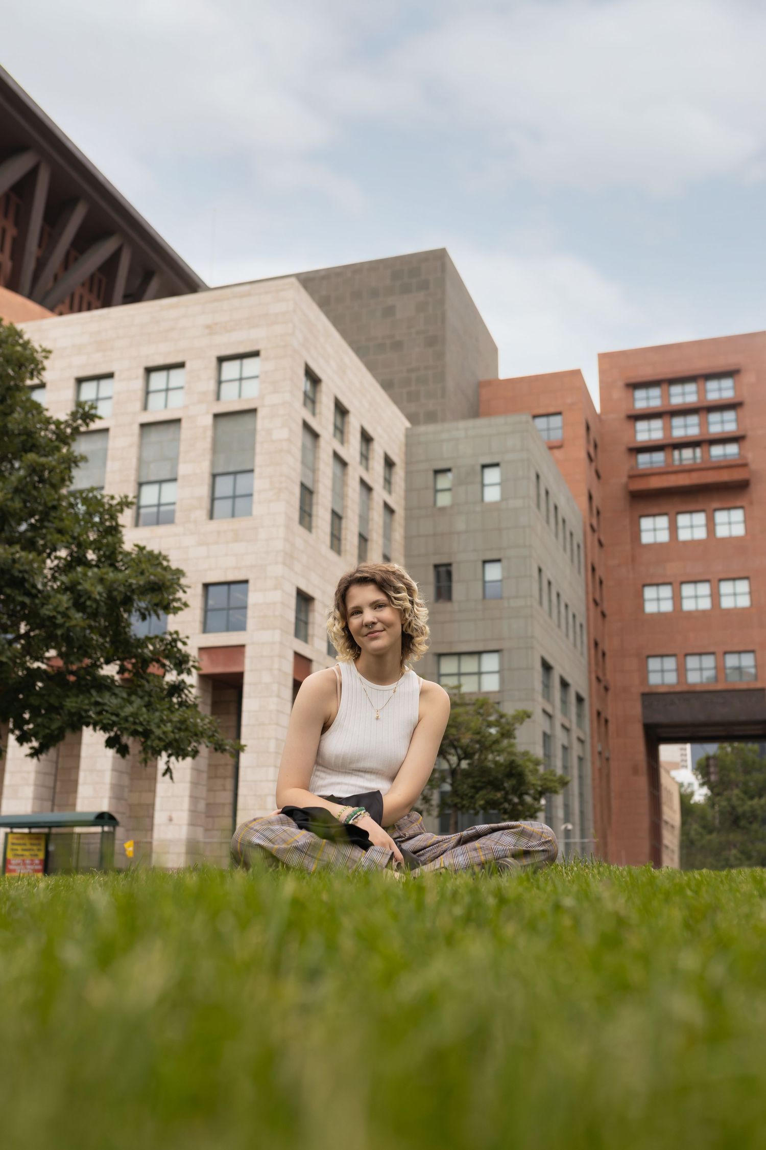 Human in white tank sitting cross-legged on grass in front of modern buildings.