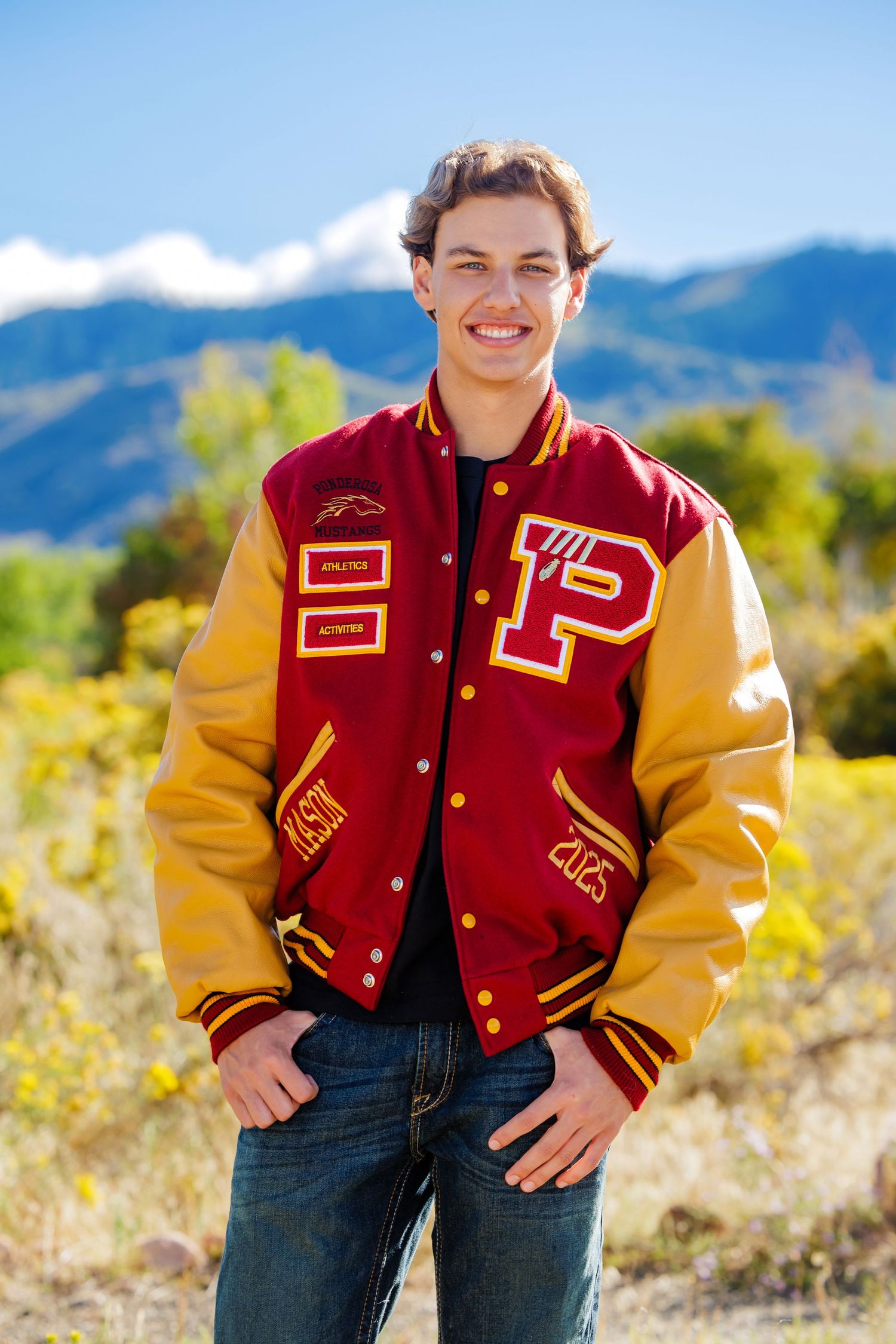 Smiling teen in red gold varsity jacket posing outdoors with mountain backdrop for senior photos.  Denver Colorado Senior Portrait