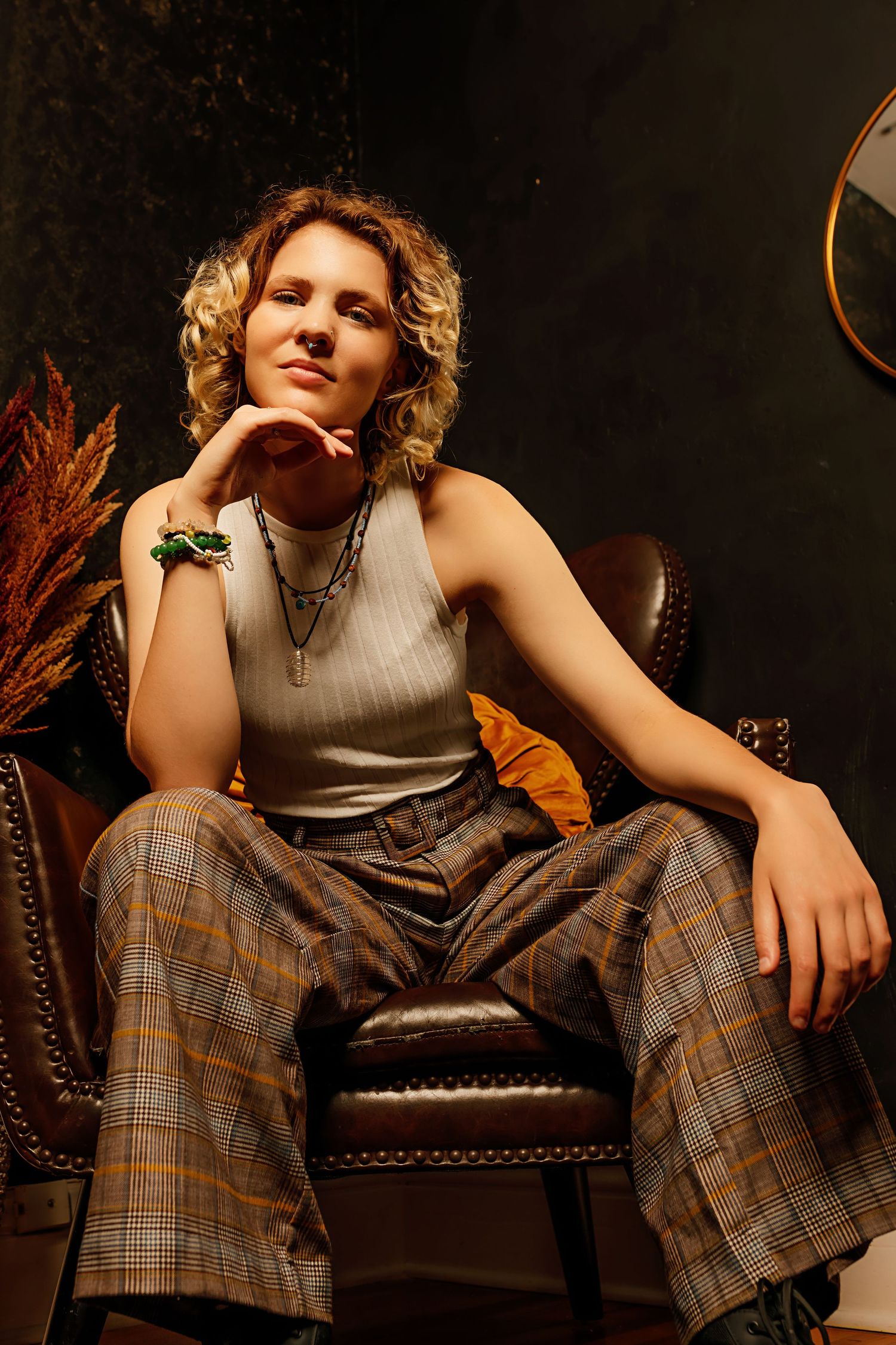 Androgynous teen in plaid flare pants and white sleeveless top poses in leather chair with green bracelet.