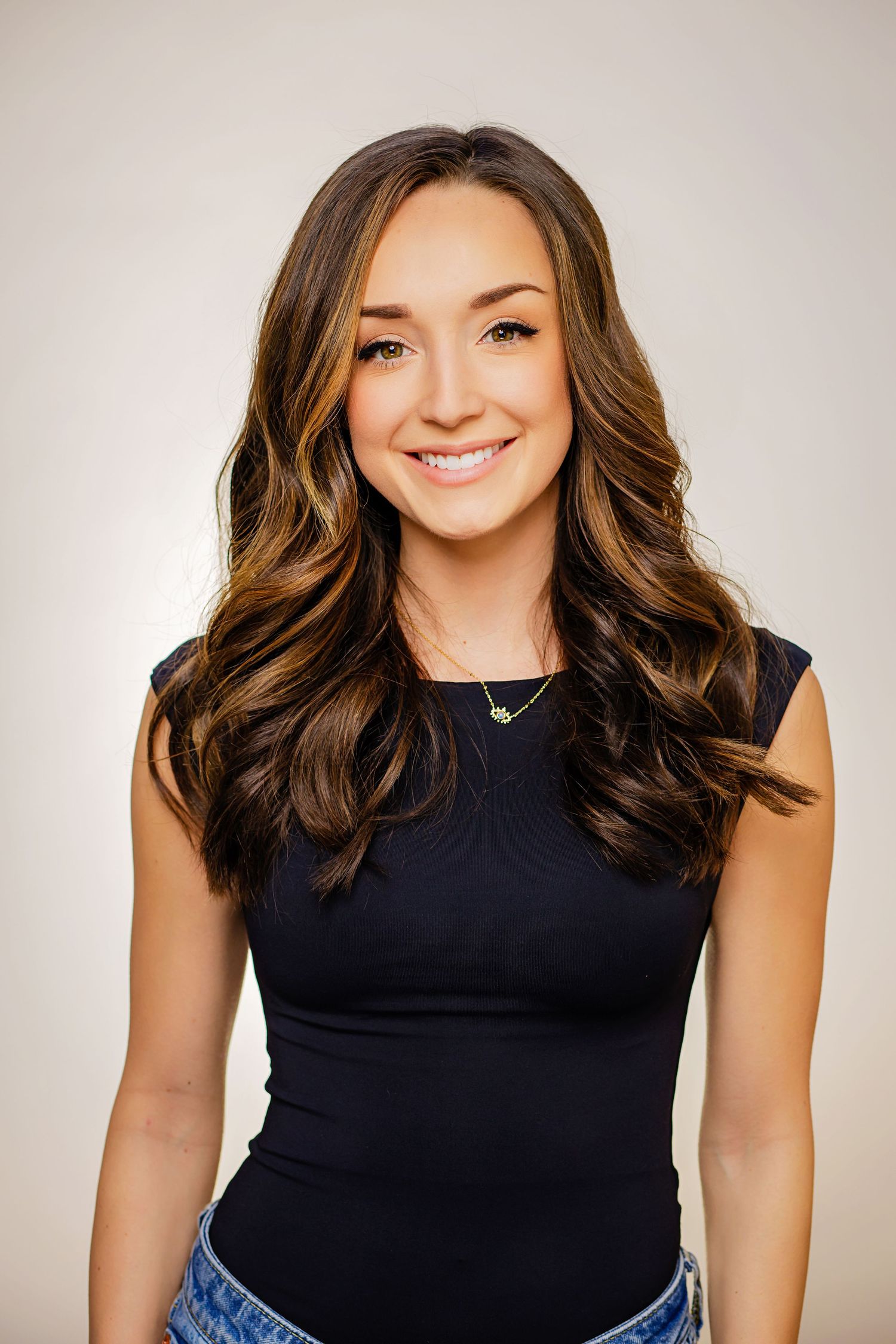 Smiling young woman with long wavy brunette hair wearing a black sleeveless top and gold necklace.