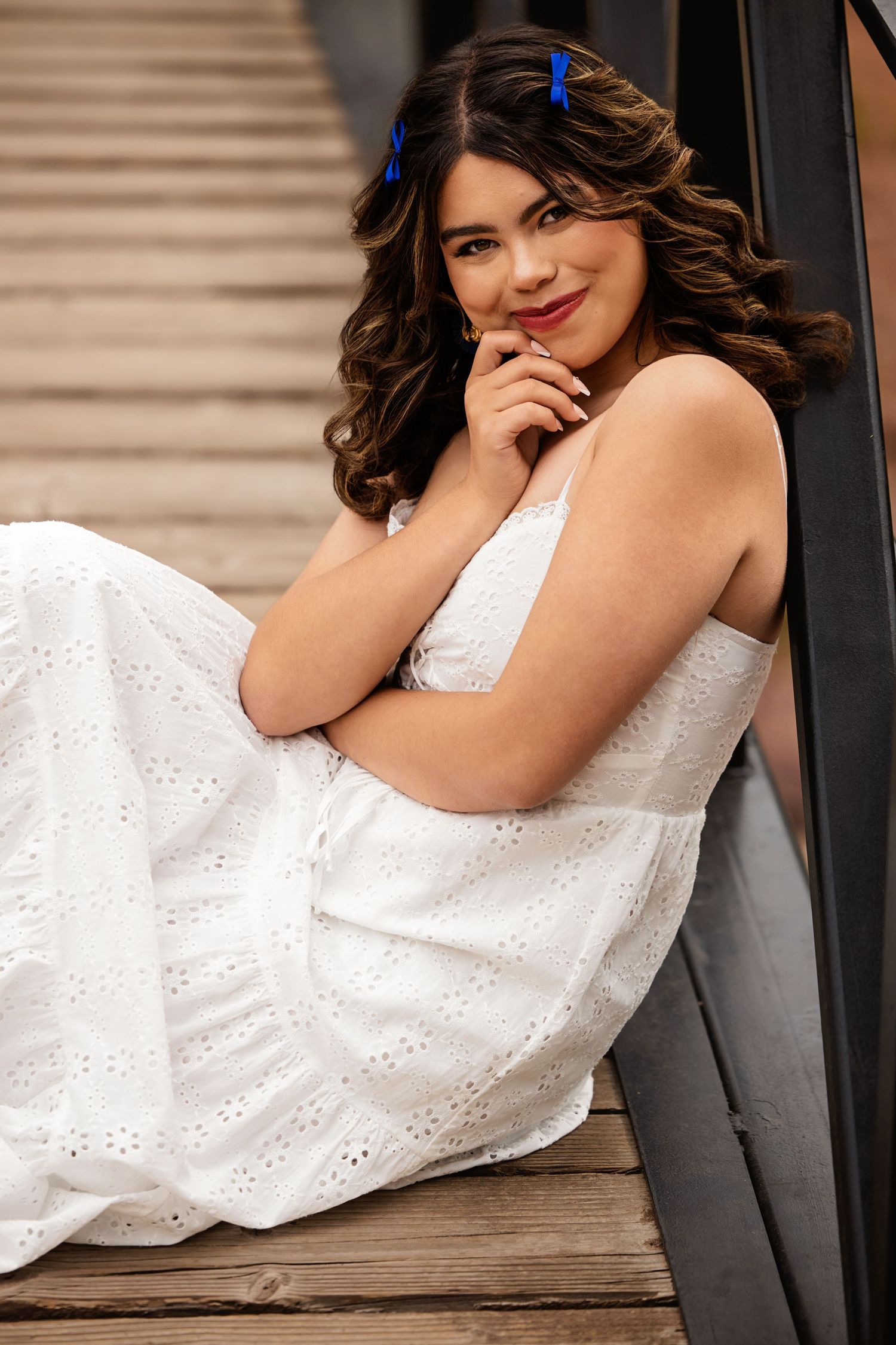 Young woman in white eyelet dress sitting on wooden steps, smiling with hand on chin, blue bow in curly hair for Colorado Senior pictures