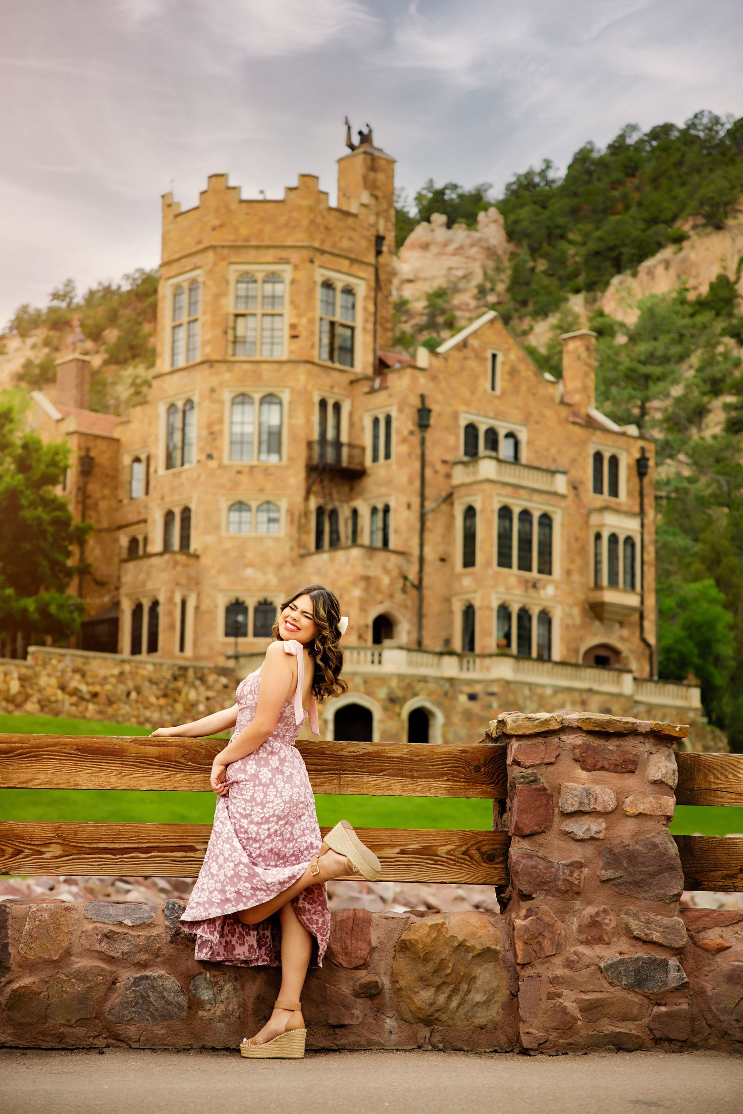 Young woman in pink floral midi dress leans against rustic fence before a grand stone castle for Aurora Colorado Senior pictures