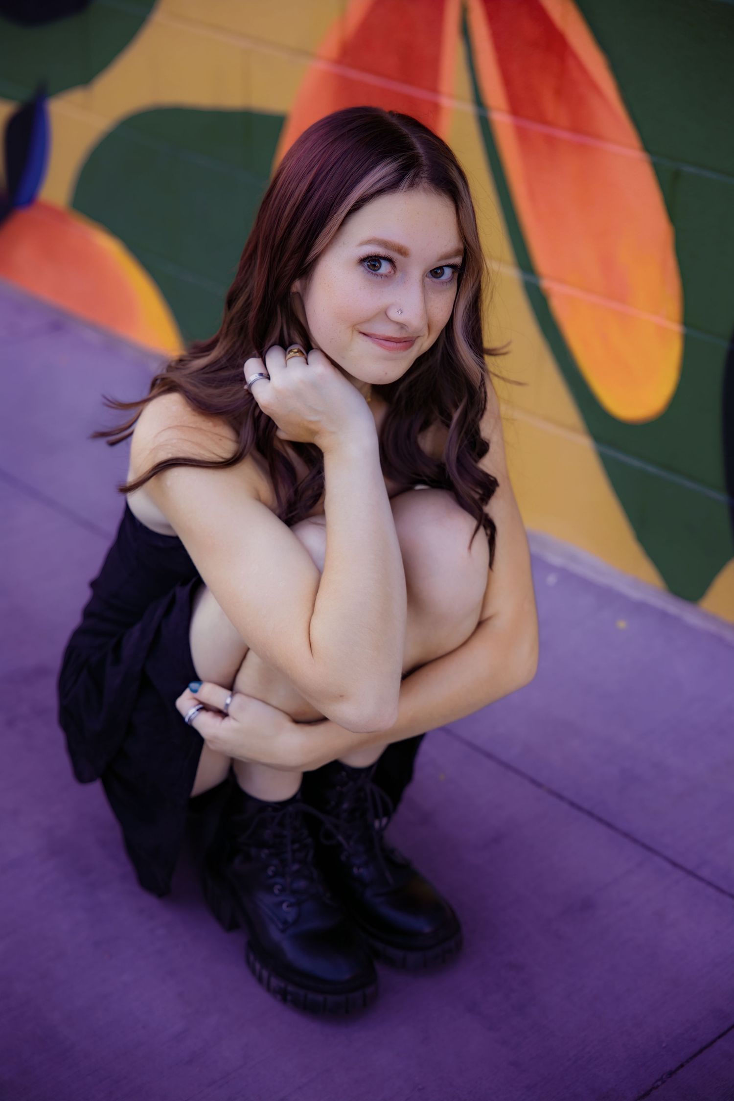 Smiling young woman in black outfit crouching against vibrant painted mural backdrop Urban Downtown Denver CO Senior photos