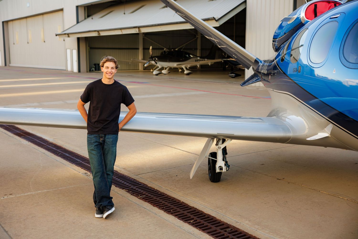 Teen boy in black shirt stands beside a blue propeller plane on an airport tarmac.