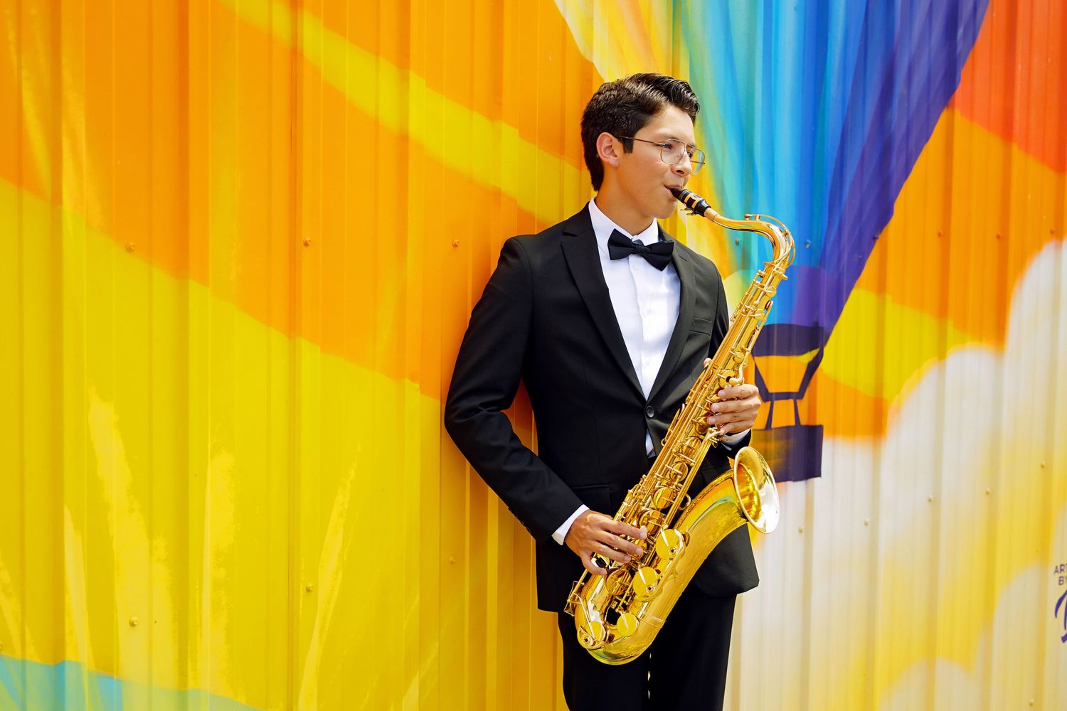Young man in tuxedo playing saxophone against colorful rainbow mural backdrop.