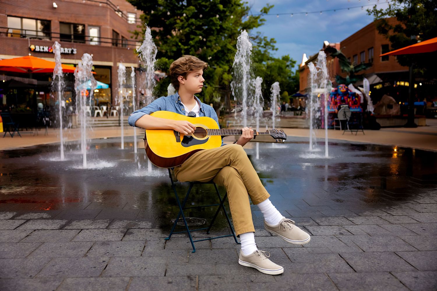 Colorado High School senior musician playing acoustic guitar seated by urban fountains on a brick plaza at dusk in